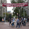 A gathering of people entering through the gates of Temple University under a red welcoming banner that reads "TEMPLE UNIVERSITY WELCOMES YOU AND YOUR FAMILY", flanked by historical stone buildings.