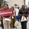 Alt-text: Two young people, the woman holding a sign with 'SAMANTHA' in white letters on a red background, lead a group of peers, all wearing lanyards and carrying bags, at an outdoor event.
